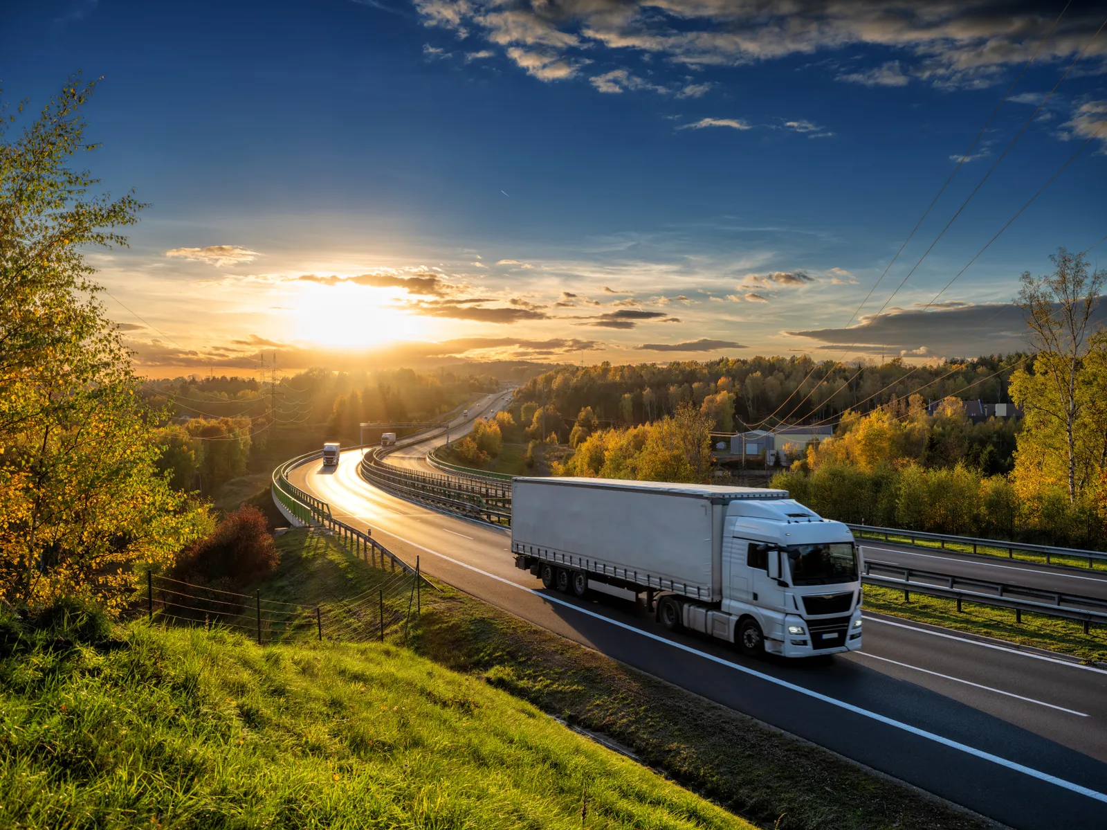 White transport trucks drive a highway winding through forested hills.