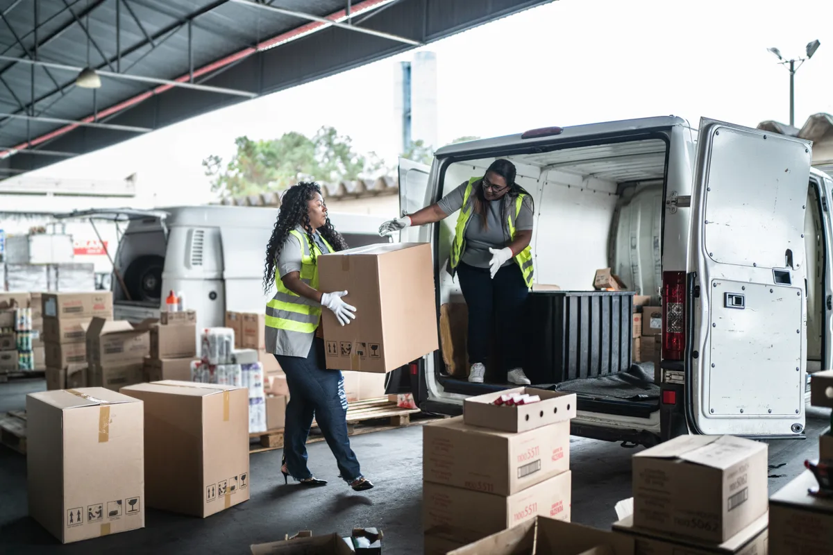 Warehouse workers in hi-vis vests load large boxes into a cargo van at a distribution centre.