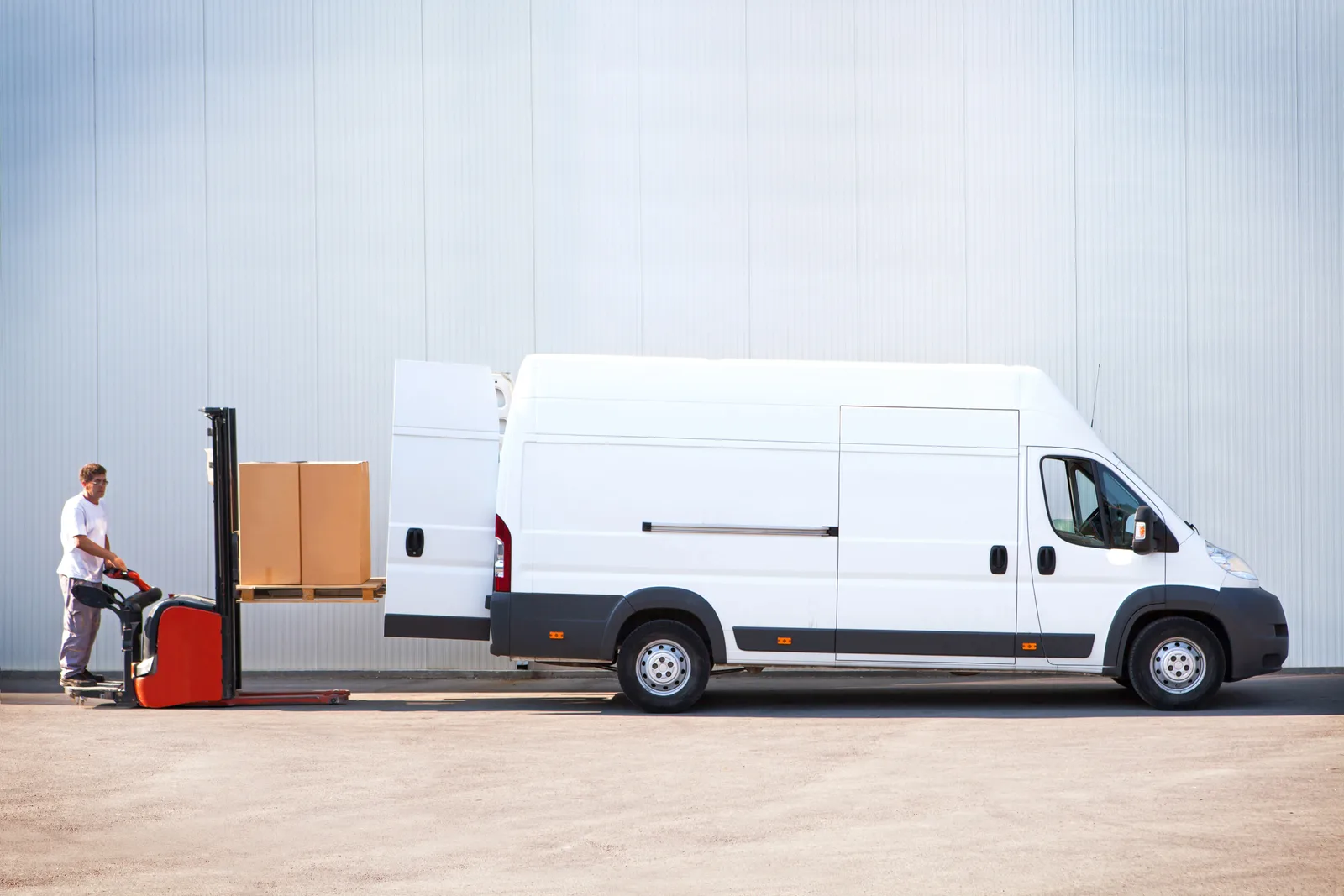 A worker loads a pallet of boxes into a white cargo van with a pallet stacker.
